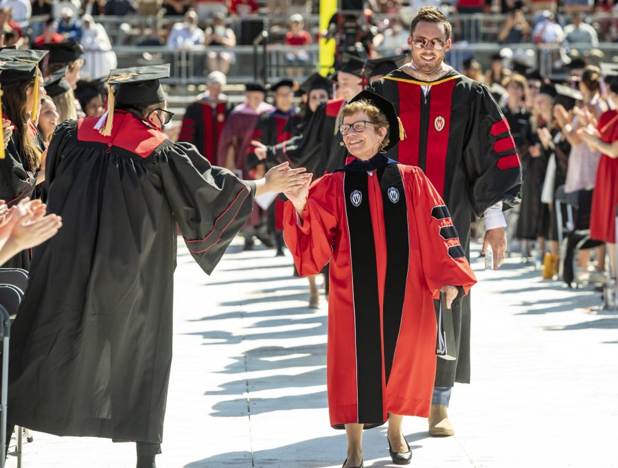 At the front of a procession of academics in regalia, Rebecca Blank extends her hand to give a high five to a graduate standing up from his chair to greet her.