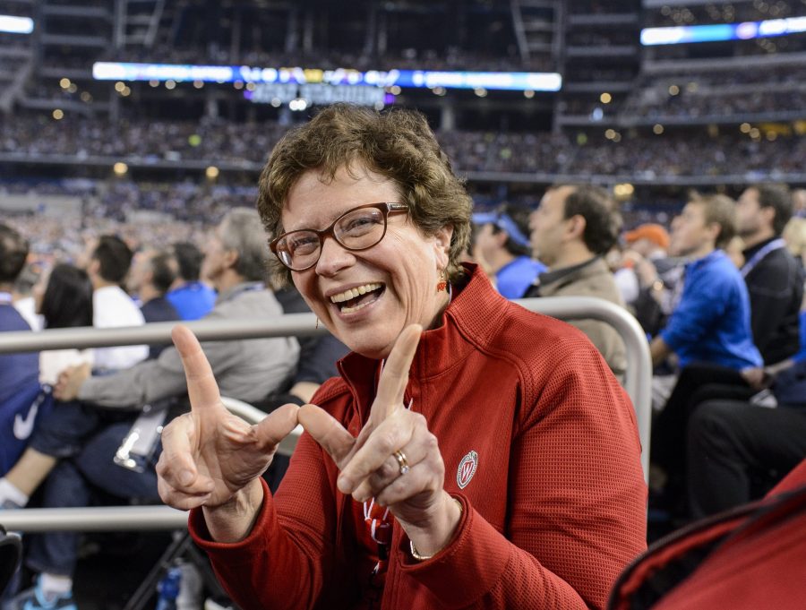 Rebecca Blank sits in the stands of the AT&T Stadium, a large indoor arena. She turns to face the camera, smiling wide and holding up her thumbs and index fingers in a W shape.