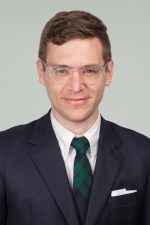 Headshot of Adam Neuman. Neuman has short brown hair and wears clear framed glasses. He is wearing a dark suit coat with a white shirt and green plaid tie.