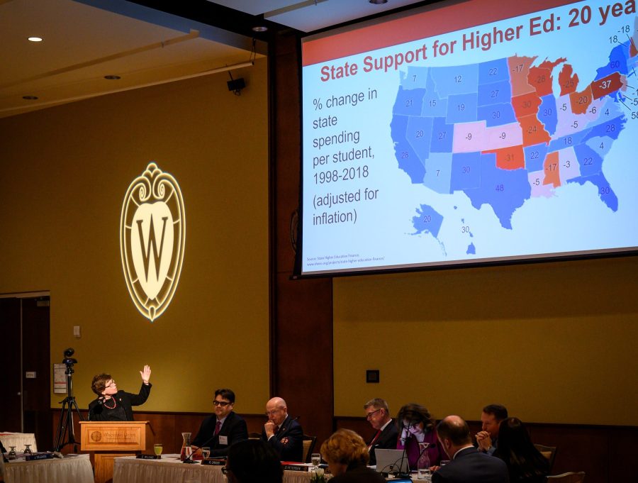 In a darkly lit banquet hall with a high ceiling, Chancellor Emerita Rebecca Blank stands at a podium and gestures up to a large projector screen. On the screen, a color-coded map of the United States shows the percent change in state support for higher education over 20 years. Wisconsin lags at negative 28% growth.