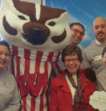 Five people father around Bucky Badger and smile. The subject of this tribute is to Bucky's left and she is wearing a red jacket and gray skirt.
