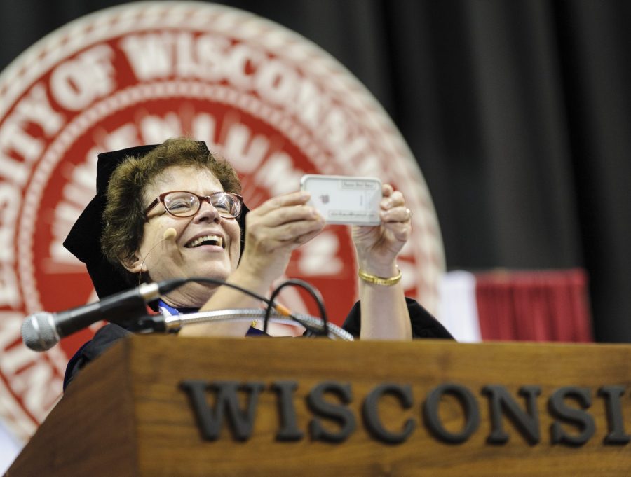 Chancellor Blanks pauses to photograph the audience with a smartphone before speaking to thousands of first-year students during the Chancellor's Convocation. She is wearing academic regalia. Behind her is the seal of UW–Madison.