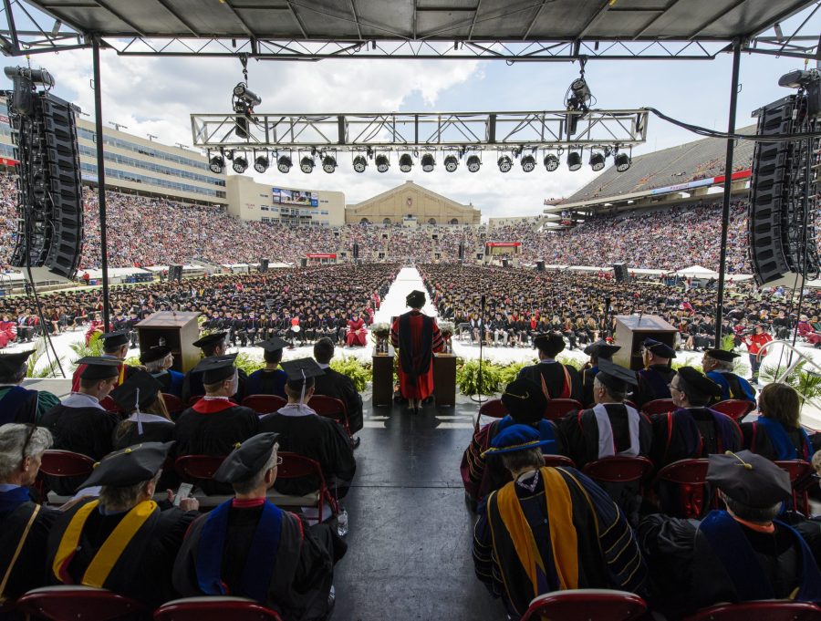 At a commencement ceremony. A wide view from the back of an elevated platform where rows of people wearing academic regalia sit facing forward listening as Rebecca Blank stands to address the crowd of graduates and their families filling Camp Randall stadium.