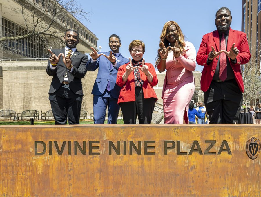 Rebecca Blank and four UW alumni, two to her left and two to her right, make a W sign with their thumbs and forefingers. They are standing just behind a newly installed marker on campus that reads 