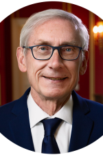A headshot of Wisconsin Governor Tony Evers. Evers is a gray-haired man. He is smiling. He wears a black suit coat and black tie and glasses.