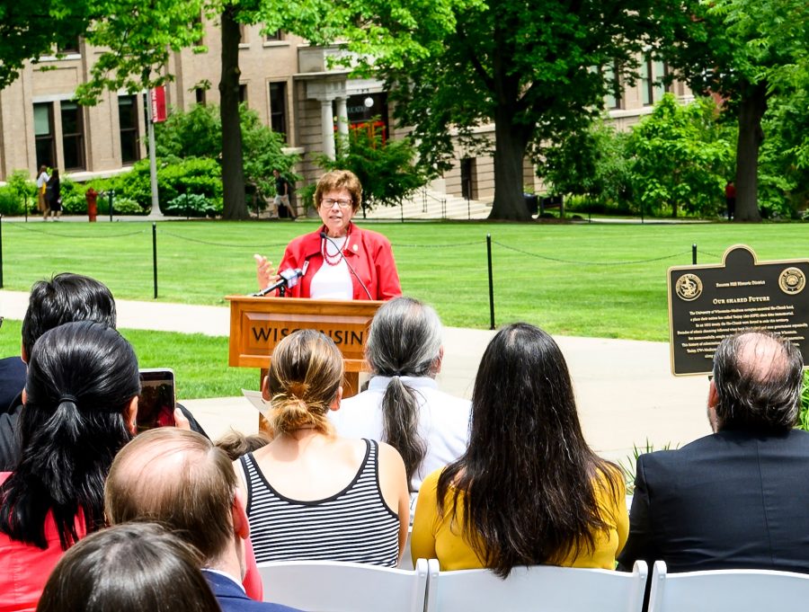 Rebecca Blank stands outdoors at the top of Bascom Hill. Behind her is a grassy lawn and the entrance to the Education Building. In the foreground, a seated crowd listens as she speaks from a lectern. The day is sunny and bright.