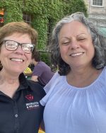 Two women smile for the camera. One, who is the subject of this tribute page, wears a black polo shirt and glasses. The other is in a white shirt.