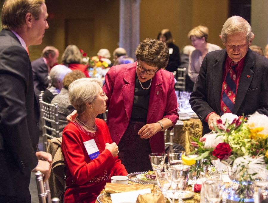 In a banquet hall, Rebecca Blank visits the table of Tashia and John Morgridge. Blank leans down to speak to Tashia, who is seated, while John stands to Blank's left. The table in the foreground is covered in fine tableware and a floral arrangement.