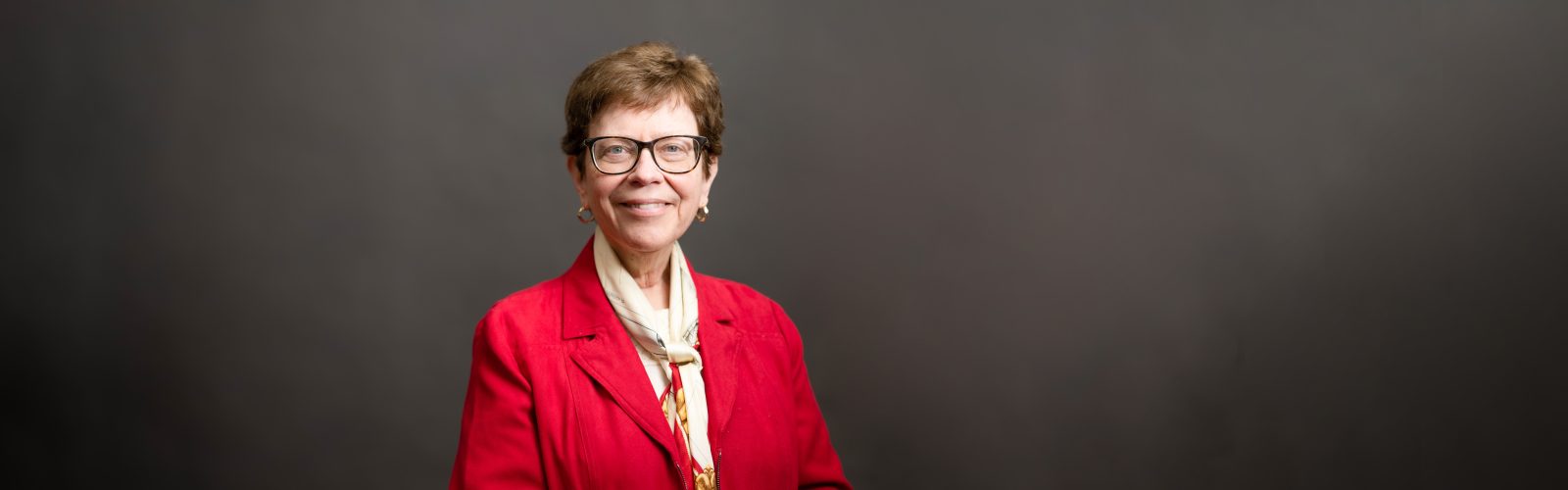 Portrait of Rebecca Blank smiling and wearing a read jacket with a white silk scarf tied loosely around her neck. She is standing against a dark gray studio backdrop.