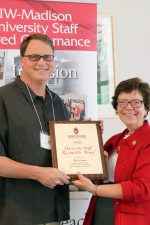 Rich Gassen and Rebecca Blank pose together for a photo holding a staff recognition award presented to Rich by Rebecca. Rich has short brown hair and is wearing a gray polo and glasses. Rebbeca has short brown hair and is wearing a red jacket with a white top, red scarf, and glasses.