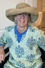 Photo of Su Hilty. Hilty sits in a wheelchair. She is wearing a brimmed straw hat and a patterned green and blue blouse.