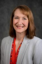 Headshot of Tammy Evetovich. Evetovich has shoulder-length auburn hair and smiles toward the camera. She is wearing a light gray suit coat and a red blouse with a colorful glass beaded necklace.