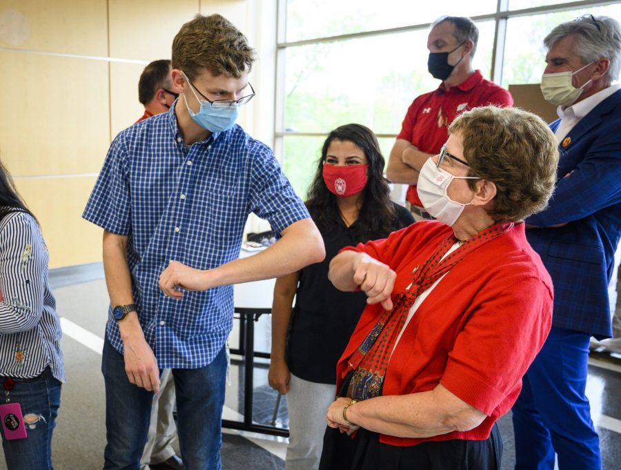 Microbiologist Jaret Schroeder elbow bumps Chancellor Blank amid a small group of people in a brightly lit indoor space. Everyone pictured is wearing surgical face masks.