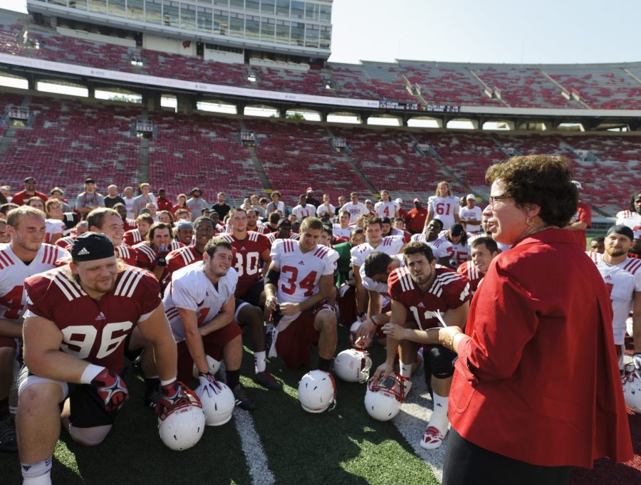 On the football field in Camp Randall Stadium, the full UW football team in game uniforms take a knee to listen as Rebecca Blank speaks to them following practice.